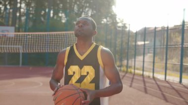 Basketbol sahasında basket oynayan yakışıklı Afrikalı basketbolcunun portresi. Spor elbiseli, kendine güvenen bir adam kameraya bakıyor. Güneşli bir gün, yaz zamanı