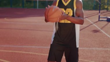 Spor giyimli yakışıklı Afrikalı bir adamın elinde topla basketbol sahasında potaya bakması ön planda. Kameraya gülümseyen yakışıklı oyuncunun portresi. Güneşli bir gün, açık havada. Sağlıklı yaşam tarzı