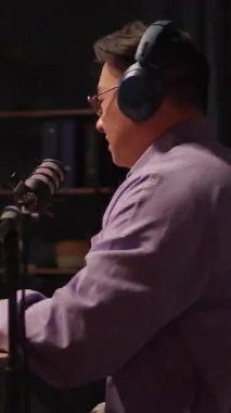 Kulaklık ve gözlüklü bir adam, mikrofon ve dizüstü bilgisayarla dolu bir masada sanatsal yaratıcılığını tutkuyla ifade ediyor ve şarkı söylüyor.