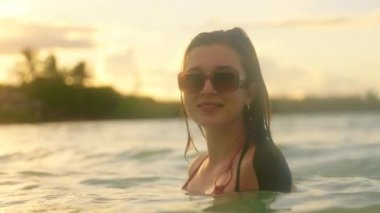 Bikinili bir kadın güneş batarken okyanustan zevk alır ve sakin bir zemin oluşturur. Sahne rahatlamayı ve mükemmel bir plaj gününün özünü yakalar.