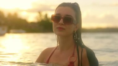 Bikinili ve güneş gözlüklü bir kadın suda duruyor, sakin bir günbatımının tadını çıkarıyor. Altın sarısı renkler, güzel bir şekilde yansıyor, somut yaz rahatlığı.