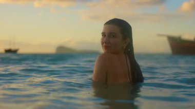 Bir kadın gün batımında sakin okyanusta zarif bir şekilde yüzer. Sıcak renkler altın bir parıltı saçar. Uzak bir tekne macerayı güçlendirir.