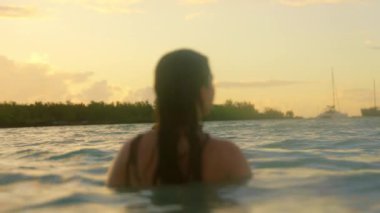 Okyanusun üzerinde sakin bir günbatımı, bir kadının suda dalgalara daldığını, teknelerin huzur içinde sürüklendiğini, doğanın rahatlığını yakaladığını gösterir.