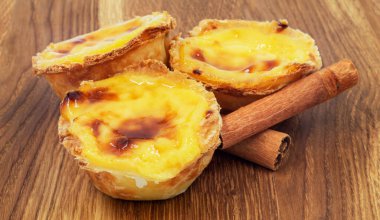 Tahta kahverengi arka planda üç Pastel de nata veya Portekiz yumurtalı tart ve tarçın çubukları. Pastel de Belem, kremalı puf böreğinden yapılan küçük bir tatlıdır.