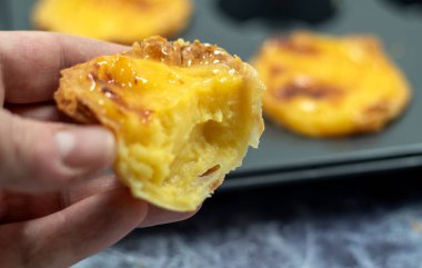 Bir adamın eli, yarısı yenmiş taze Portekiz Pastel de Nata turtasının arkasındaysa muhallebi. Pastel de Belem yanmış kapağı ve çıtır börek hamuru olan bir hamur işidir.
