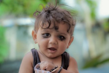 Kıvırcık kahverengi saçlı, kameraya gülümseyen genç bir Hintli çocuk. Bebek kolsuz siyah bir gömlek ve altın bir bilezik takıyor..