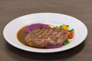 Beef steak on a white plate with grilled tomatoes, asparagus, mashed purple sweet potato and brown sauce.