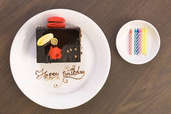 Birthday chocolate cake decorated with macarons and white chocolate on a white plate with the words Happy Birthday.