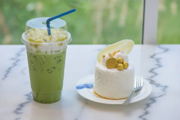 Ice matcha green tea in plastic cup and Coconut cake decorated with white chocolate, biscuits and gold leaf in a white plate on a granite table with a garden background.