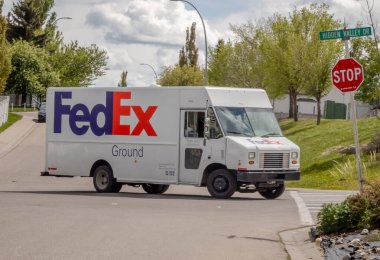 Calgary, Alberta, Kanada. 4 Haziran 2024.A FedEx arazi kamyonu dur işareti ve sokak tabelasıyla işaretlenmiş bir kavşakta dönüyor, yemyeşil ve parçalı bulutlu bir gökyüzü olan bir yerleşim bölgesinde..