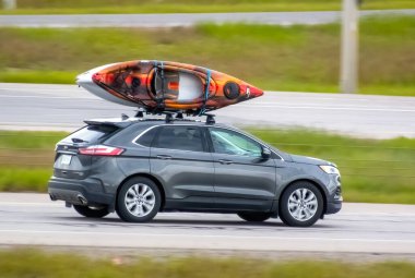 Calgary, Alberta, Kanada. 21 Haziran 2024. Gri bir Ford Edge, siyah çatı rafına monte edilmiş turuncu ve beyaz bir kanoyla otoyolda ilerliyor..