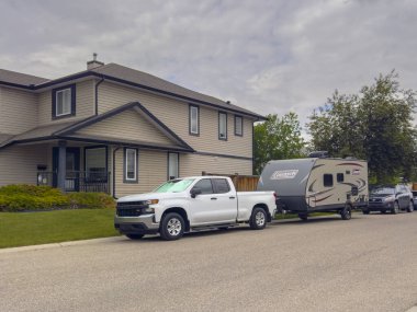 Calgary, Alberta, Kanada. 19 Haziran 2024. Park etmiş beyaz bir kamyonet Coleman karavanı çekiyor. İki katlı, bej kaplamalı modern bir evin önünde..