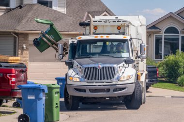 Calgary, Alberta, Kanada. 25 Haziran 2024. Mekanik kolu olan beyaz bir çöp kamyonu evlerle dolu bir sokakta çöp tenekesini kaldırıyor. Olay yeri tipik bir çöp toplama sürecini betimliyor.