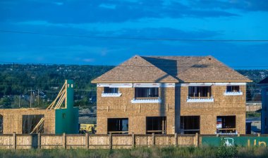 Calgary, AB, Canada. Aug 27, 2024. A two-story house under construction with a wood frame and plywood sheathing. The house is surrounded by a wooden fence and has a view of a nearby community. clipart