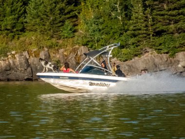 Waterton Park, AB, Kanada. Ağustos 24, 2024. Bir Malibu teknesi gölde hızla ilerlerken ardında iz bırakır. Yolcular yolculuğun tadını çıkarırken, bir köpek kayalık bir uçurumdan izliyor..