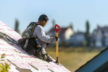 Calgary, AB, Kanada. 19 Eylül 2024. Bir inşaat işçisi eğimli bir çatıda çatıdaki küreği dikkatlice kullanıyor, açık gökyüzünün altında güvenlik donanımı giyiyor. Endüstriyel çatı küreği.