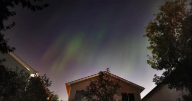 Aurora Borealis manzarası banliyö evlerinin üstündeki gece gökyüzünü aydınlatır, aşağıdaki ağaçlar ve çatılar üzerinde ruhani bir parıltı bırakır..