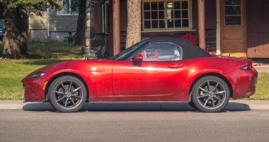 Waterton Park, AB, Canada. Aug 20, 2024. A vibrant red Mazda MX-5 Miata convertible gleams in the sunlight, parked alongside a charming wooden house with a backdrop of lush green trees. clipart