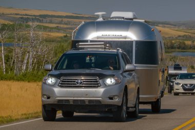 Waterton Park, AB, Kanada. 19 Ağustos 2024. Gümüş renkli bir Toyota Highlander SUV, arka planda kayan tepeler ve uzak bir göl bulunan, manzaralı bir otoyol boyunca klasik bir Airstream römorku çekiyor..