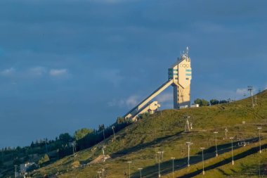 Calgary, AB, Canada. Aug 27, 2024. A Canada Olympic Park ski jump tower stands tall against a blue sky, surrounded by green hills and a chairlift. The tower has a distinctive Olympic rings symbol. clipart