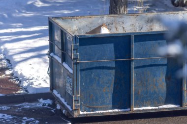 Calgary, Alberta, Kanada. 23 Ekim 2024. Metal kapaklı mavi bir çöp konteynırı kısmen karla kaplıdır. Çöp bidonu kaldırımda duruyor. Etrafı karla kaplı..