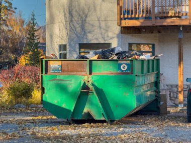 Calgary, Alberta, Kanada. 23 Ekim 2024. Evin önüne park edilmiş çöplerle dolu yeşil bir çöp konteynırı. Çöp konteynırı bir evin önündeki yapraklarla çevrili..