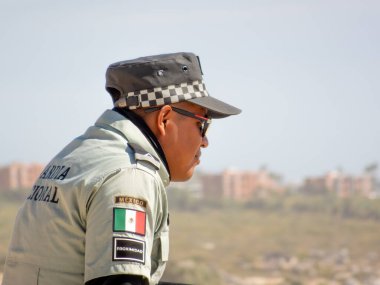 Los Cabos, B.C.S., Meksika. 26 Kasım 2024. Meksika 'dan gelen, güneş gözlüğü takan ve milli bayrak bantlı üniformalı bir polis memuru, düşünceli bir şekilde uzaklardan bakıyor..