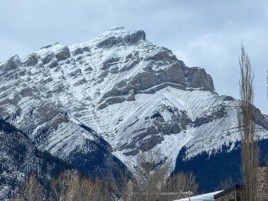 The majestic Cascade Mountain stands tall, its peak dusted with snow against a cloudy sky. Located in the Bow River Valley of Banff National Park, adjacent to the town of Banff. clipart