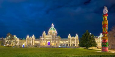 Victoria, British Columbia, Canada. Feb 9, 2025. Victoria's BC Parliament Buildings glow with festive lights against a deep blue evening sky, complemented by a vibrant totem pole on the side. clipart