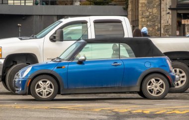 Victoria, British Columbia, Kanada. 15 Şubat 2025. Üstü açık mavi bir Mini Cooper iki büyük beyaz kamyonun arasına park etmiş..
