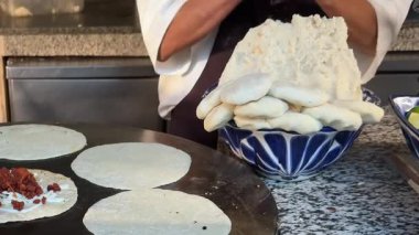Bir şef, Meksika usulü mısır tortillalarını mavi desenli bir kasede pişmiş tortilla ile bir tepsi hamurdan hazırlıyor ve bir ızgara ile dolduruyor. Aşçılık hazırlığı devam ediyor.