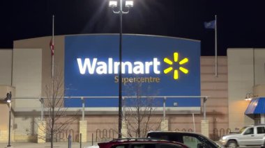 Calgary, AB, Kanada, 20-25 arası. Gece vakti aydınlık mavi ve sarı ışıklarıyla karanlık gökyüzüne karşı duran bir Walmart Supercenter. Arabalar park yerinde..