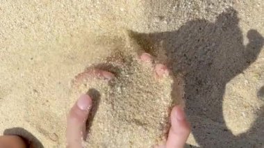 Elleri hafifçe açık renkli kumsaldaki kumu kavrıyor. Dokusunu ve güneşli bir günün sıcaklığını sergiliyor. Basit bir anda elde edilen doğal, dokunsal bir deneyim..