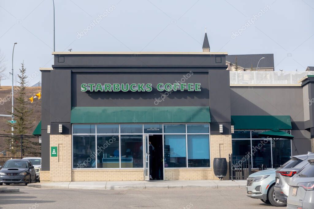 Calgary, Alberta, Canada. Mar 23, 2025. A street-level shot of a Starbucks Coffee outlet with a dark facade, featuring its logo and entrance, situated in a parking lot with cars