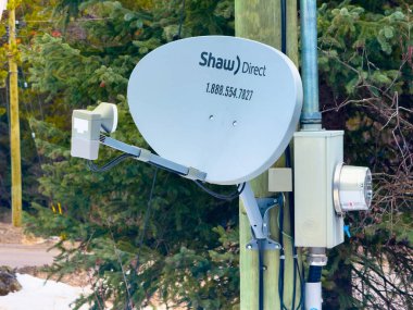 Panorama, British Columbia, Kanada. 31 Aralık 2025. Tahta elektrik direğine bağlanmış bir Shaw Direct uydu anteni. Kablolar ve bir de sayaç kutusu var. Evergreen 'ler ve caddeler bg' de.