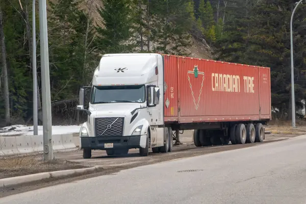 Radyum, British Columbia, Kanada. 31 Aralık 2025. Kırmızı Kanada lastikli beyaz bir Volvo yarı kamyon kaldırımlı bir yol boyunca ilerliyor, ağaçlar ve yamaçta bir yamaç var..