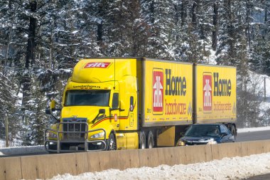 Banff, Alberta, Kanada. 2 Nisan 2025. Parlak sarı bir ev donanımı yarı kamyon, karlı bir otoyolda gidiyor, yanında bir araba, bir orman zemininin ortasında..