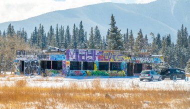 Canmore, Alberta, Kanada. 2 Nisan 2025. Grafiti ile kaplı terk edilmiş bir bina karlı bir arazide duruyor. Arkasında dağlar ve ağaçlar var. Park halindeki iki arabanın yanında..