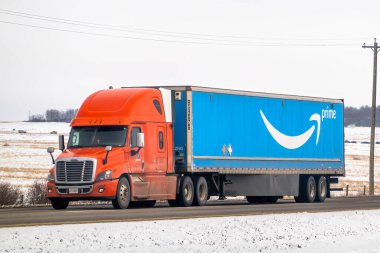 Banff, Alberta, Kanada. 2 Nisan 2025. Turuncu bir Amazon yarı kamyoneti, arka planda elektrik direği olan kırsal bir alanda karlı bir yolda ilerliyor..