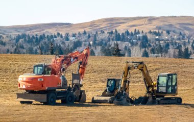 Calgary, Alberta, Kanada. 5 Nisan 2025. İki kazıcı, biri turuncu, biri sarı, açık bir gökyüzünün altında park halinde. Arkamızda bir tepe ve ağaçlar var..
