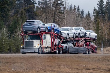 Calgary, Alberta, Kanada. 5 Nisan 2025. Çok katlı araç nakliyecisi, ağaçlık bir otoyol boyunca hareket halindeki otomotiv lojistiklerini gösteren birkaç yeni araç taşıyor..