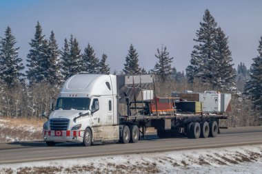 Calgary, Alberta, Kanada. 4 Nisan 2025. Ulusal Flatbed Solutions LLC tarafından işletilen beyaz bir yarı kamyon, kar boyunca bir karavanda çeşitli örtülü ve üstü açık eşyalar taşıyor..