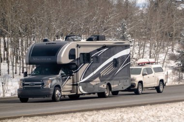 Banff, Alberta, Kanada. 4 Nisan 2025. Bir 2023 Thor Magnitude RS36 karavanı, beyaz bir kamyoneti çatı rafında eşyalarla birlikte çekiyor. Çevre karlı görünüyor.,