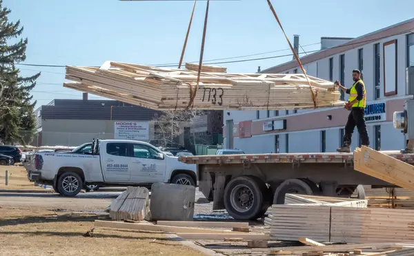 Calgary, Alberta, Kanada. 5 Nisan 2025. Vinç, güneşli bir inşaat sahasında, bir işçi tarafından düz bir kamyona yönlendirilen prefabrik ahşap çatı kirişlerini kaldırıyor..