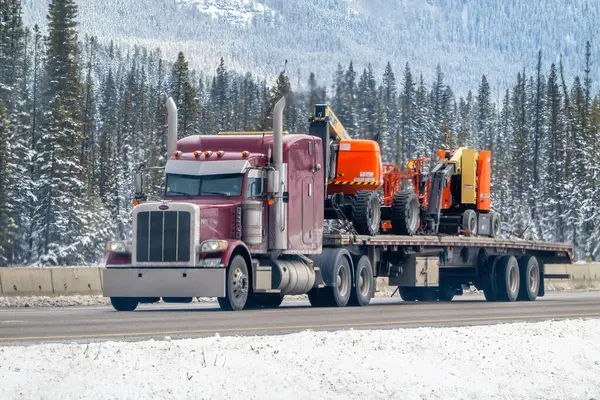 Banff, Alberta, Kanada. 4 Nisan 2025. Bir kestane rengi yarı kamyon Alberta 'nın dağlık, ormanlı bir bölgesinde karlı bir otoyol boyunca iki adet turuncu JLG patlayıcı taşıyıcı taşıyor..