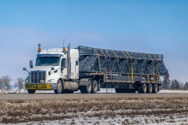 Banff, Alberta, Kanada. 4 Nisan 2025. Düz bir karavanda, büyük ihtimalle açık bir gökyüzünün altındaki kırsal ve muhtemelen karlı bir alanda, bir otobanda büyük miktarda koyu metal yapılar taşıyan beyaz bir kamyonet..