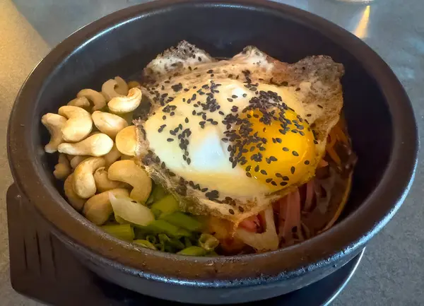 Bir taş kase bibibimbap, Kore karışık pirinç yemeği, üzerinde siyah susam tohumları bulunan kızarmış yumurta, sebze, et ve kaju fıstığı içerir..