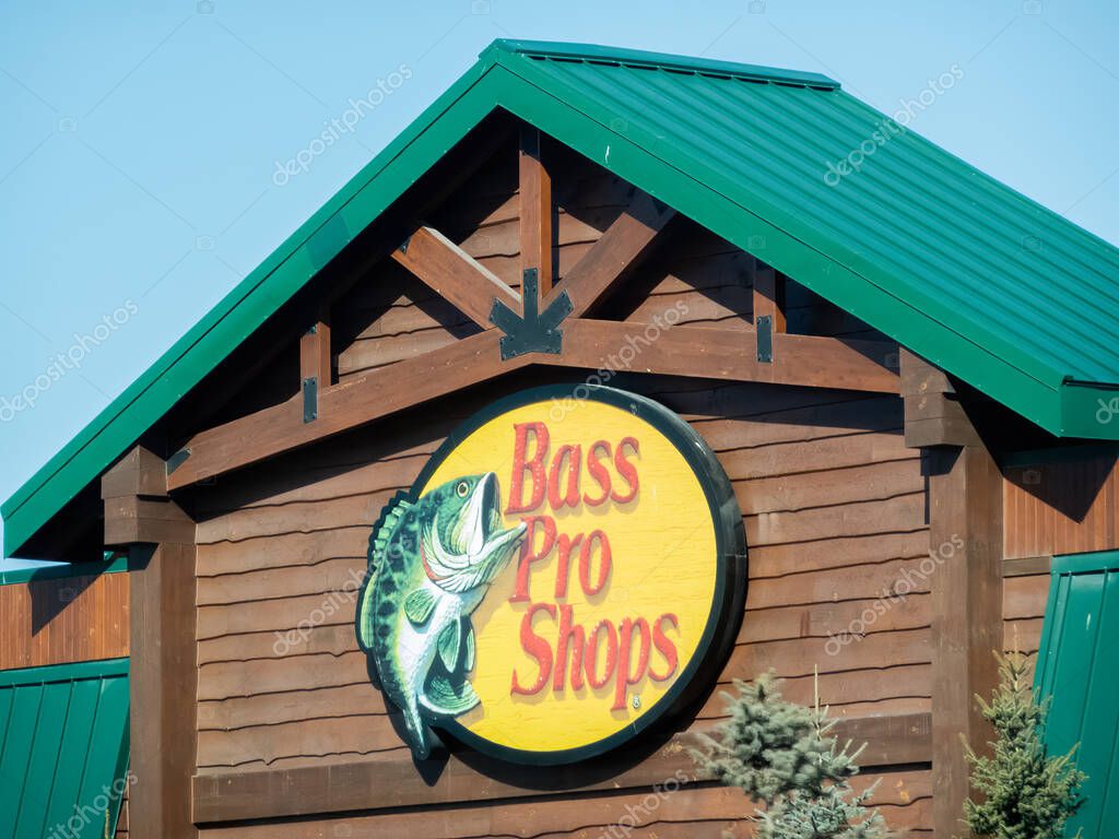 Calgary, Alberta, Canada. Apr 5, 2025. The iconic Bass Pro Shops logo adorns the rustic wood facade of a retail building with a green metal roof, under a clear sky.