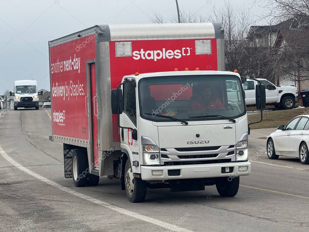 Calgary, Alberta, Canada. Apr 4, 2025. A Staples delivery truck, an Isuzu NPR, drives on a road in Calgary, Alberta, with other vehicles visible on an overcast day, suggesting a routine delivery.