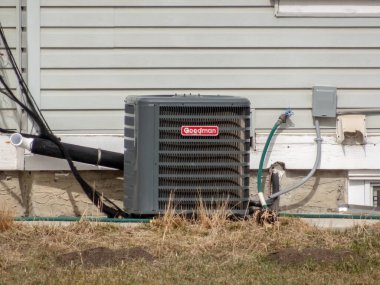 Calgary, Alberta, Kanada. 8 Nisan 2025. Goodman marka bir klima sistemi evin dışında, ışık yalıtımlı, hortumlara ve elektrik kablolarına bağlı, kuru otlarla çevrili bir evde oturuyor..